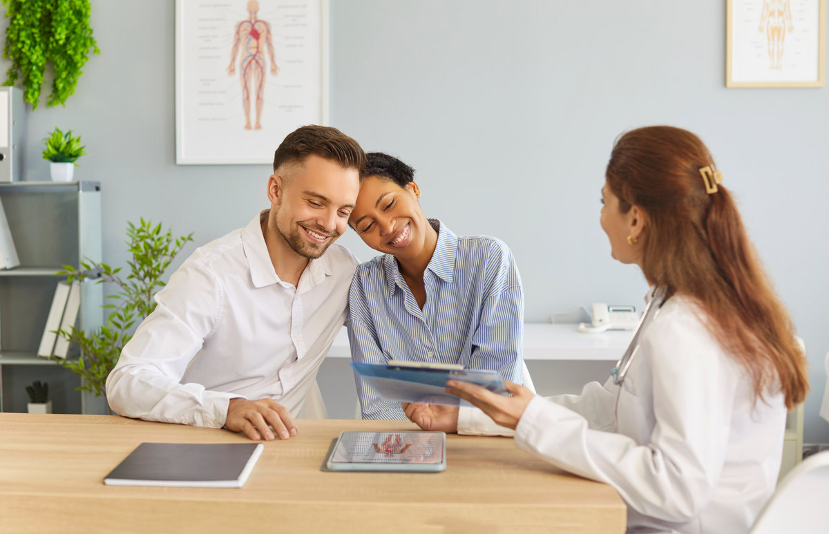 Happy young couple sitting at desk and talking with family doctor therapist looking to report file with appointment during medical exam in clinic planning pregnancy. Health care concept.