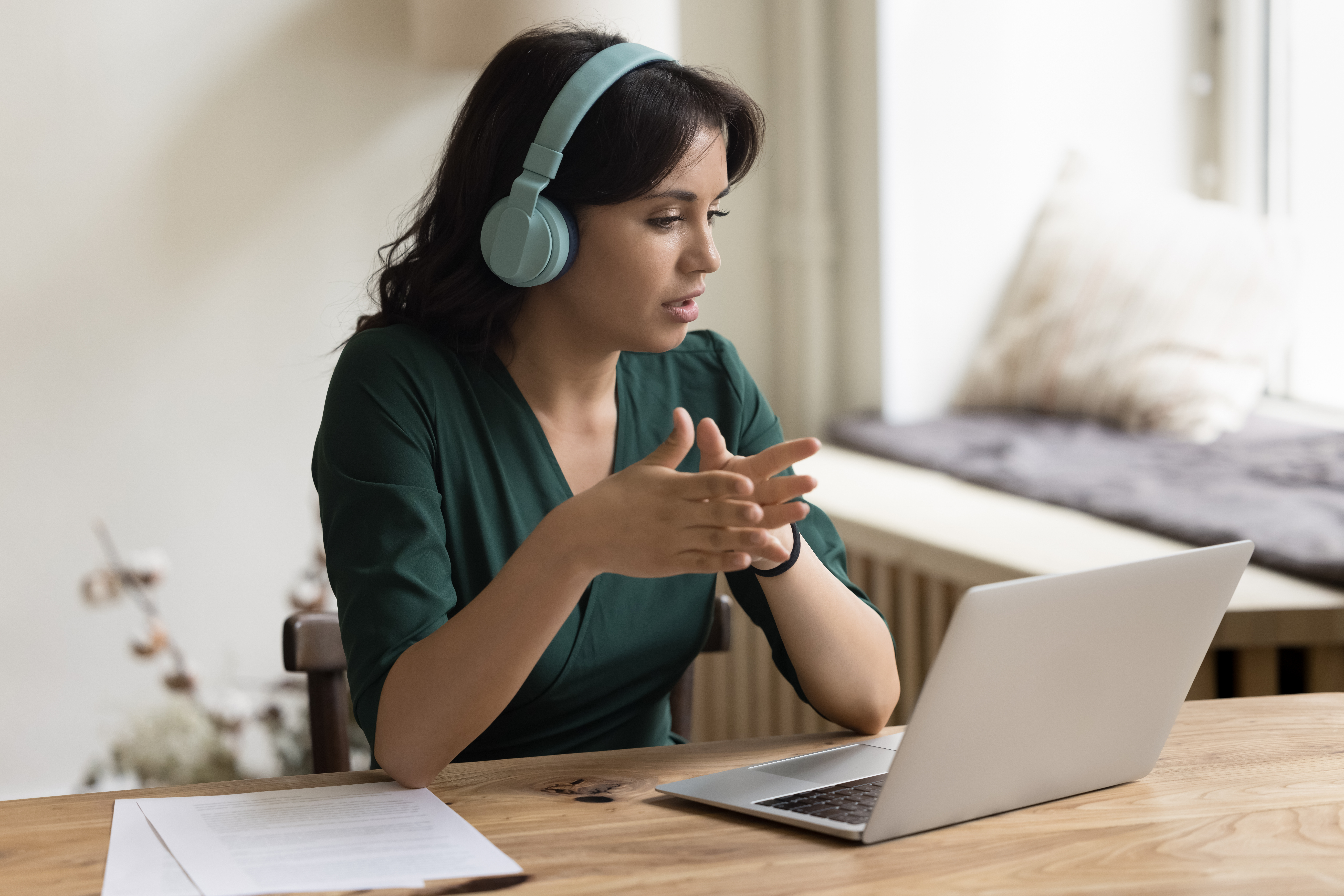 Serious Asian freelance worker woman in trendy wireless headphones talking on video call, using laptop computer for online business communication with client, speaking on Internet conference